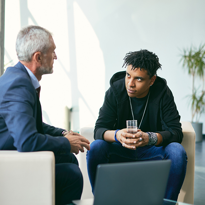 Two men engaged in a conversation in a modern office setting, one holding a glass of water while listening attentively to the other. The scene conveys a professional dialogue focused on important matters.