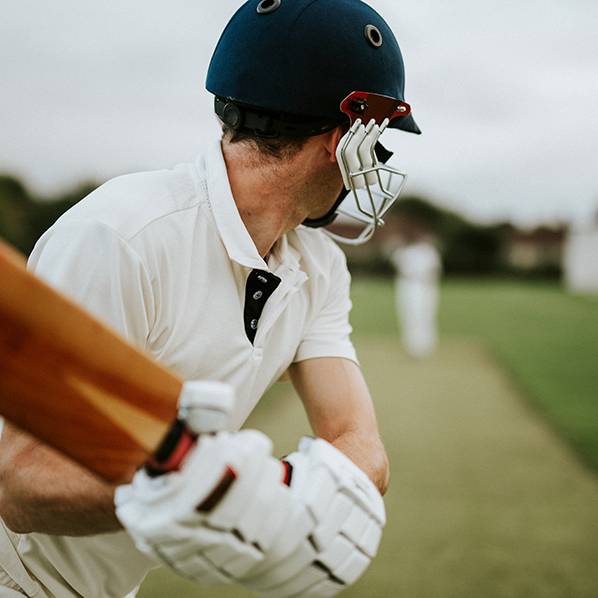 Cricketer preparing to bat on a green pitch, wearing a helmet and holding a wooden bat.