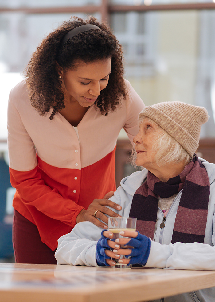 A caring young woman leans towards an elderly woman, offering comfort and companionship as they share a moment over a drink in a cozy setting.