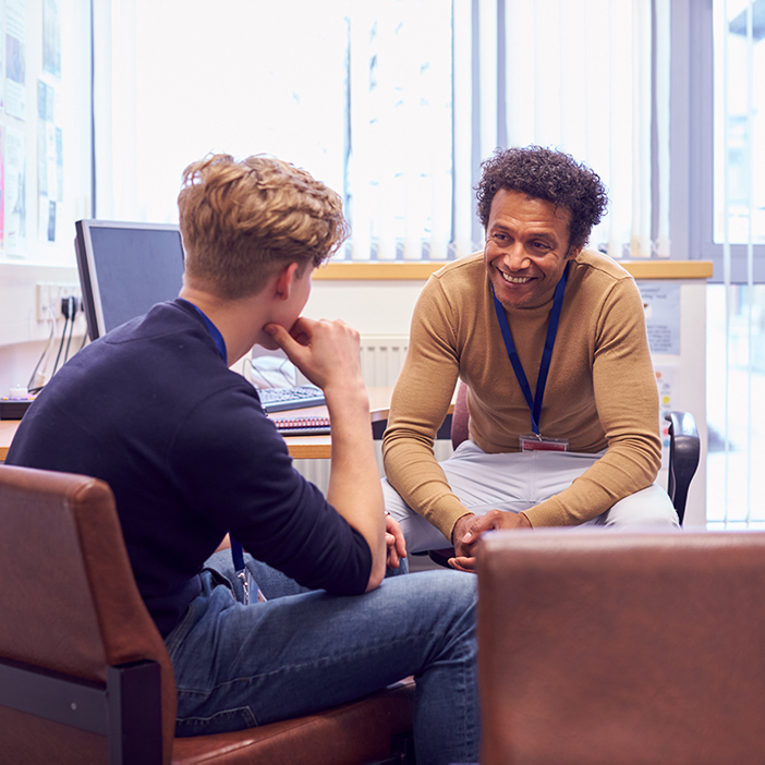 A smiling man in a beige sweater talks to a young man seated in an office setting, fostering discussion and support.